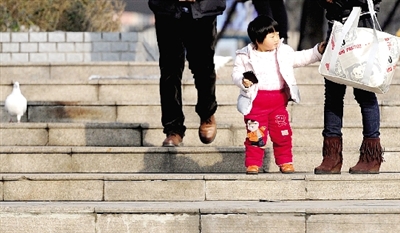 昨天中午，一女孩拉著家長的手在紫荊山公園曬太陽，氣溫升高，孩子的棉衣解開了扣子。
